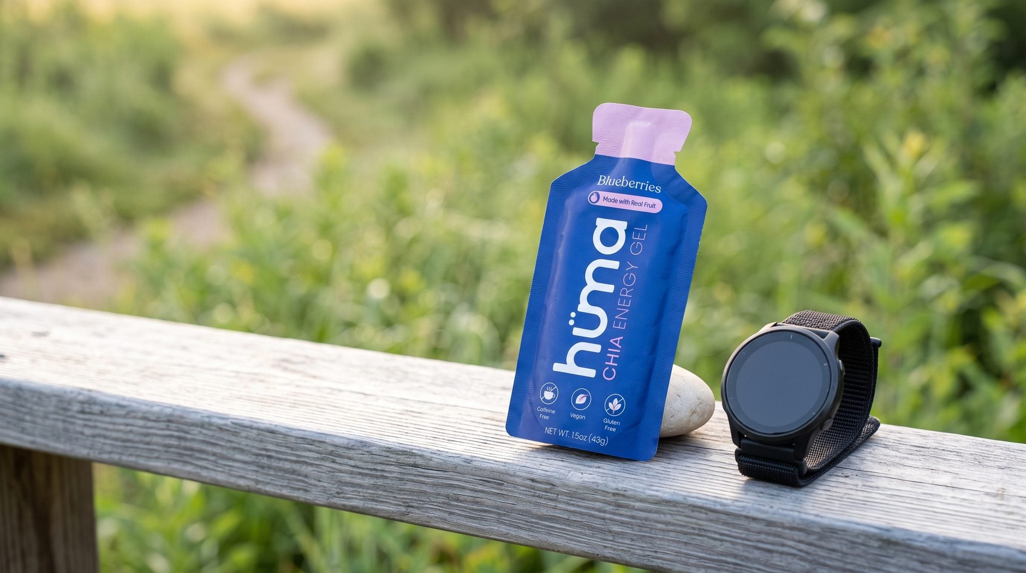 A Huma Blueberries chia energy gel leaning against a stone next to a GPS running watch on a wooden rail at a sunrise trailhead