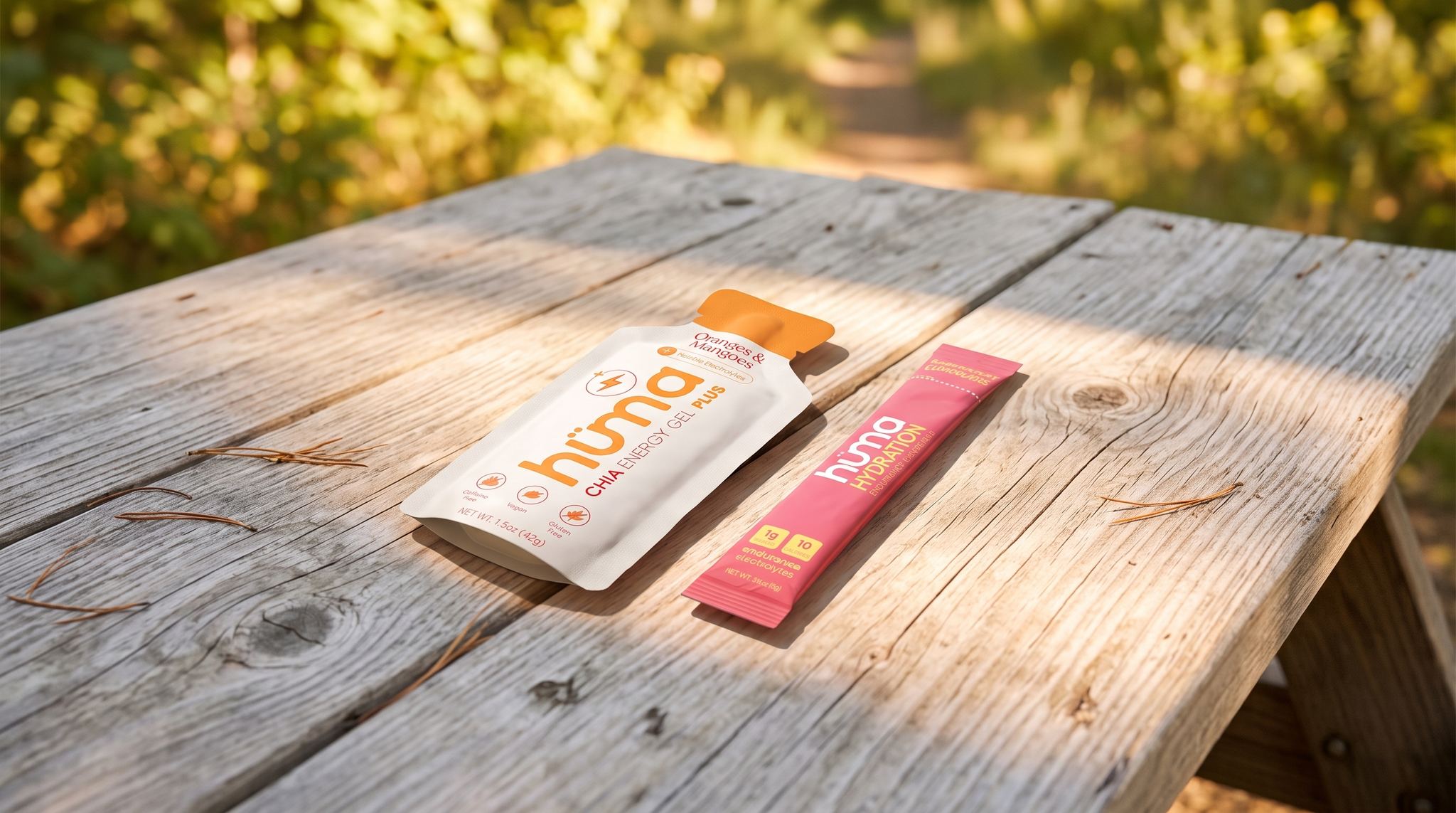 A Huma PLUS Oranges and Mangoes gel and a pink Huma Hydration Raspberry Lemonade stick pack resting on a weathered wooden picnic table in dappled summer sunlight at a trailhead
